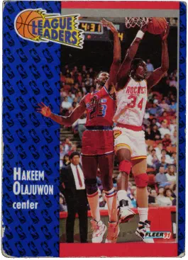 The front of a 1991 Fleer basketball card featuring Hakeem Olajuwon. In the photo Hakeem has caught a rebound while an opposing player behind him has failed.